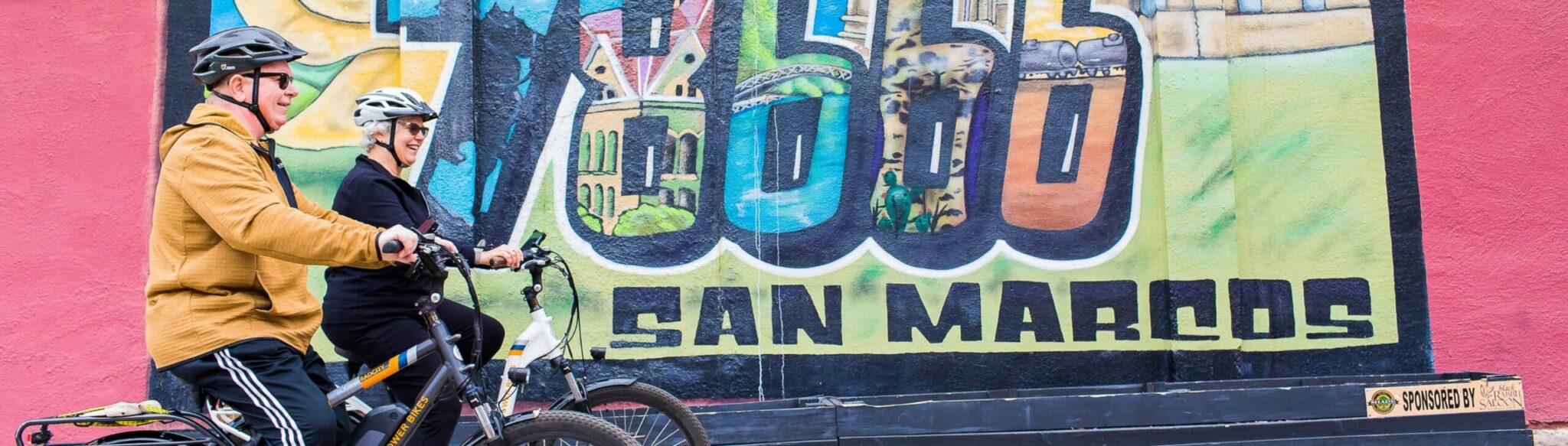 78666 San Marcos mural on building with couple riding bikes in front